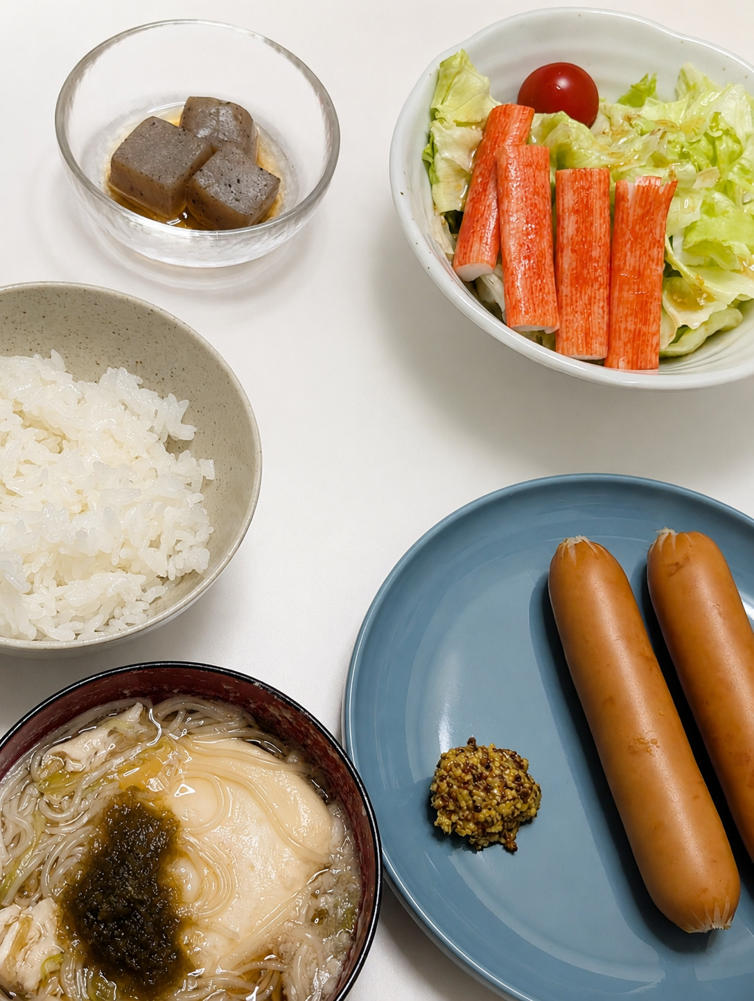 こんにゃく煮物・カニカマサラダ・白米・ソーセージ・汁物が並んだ夕食