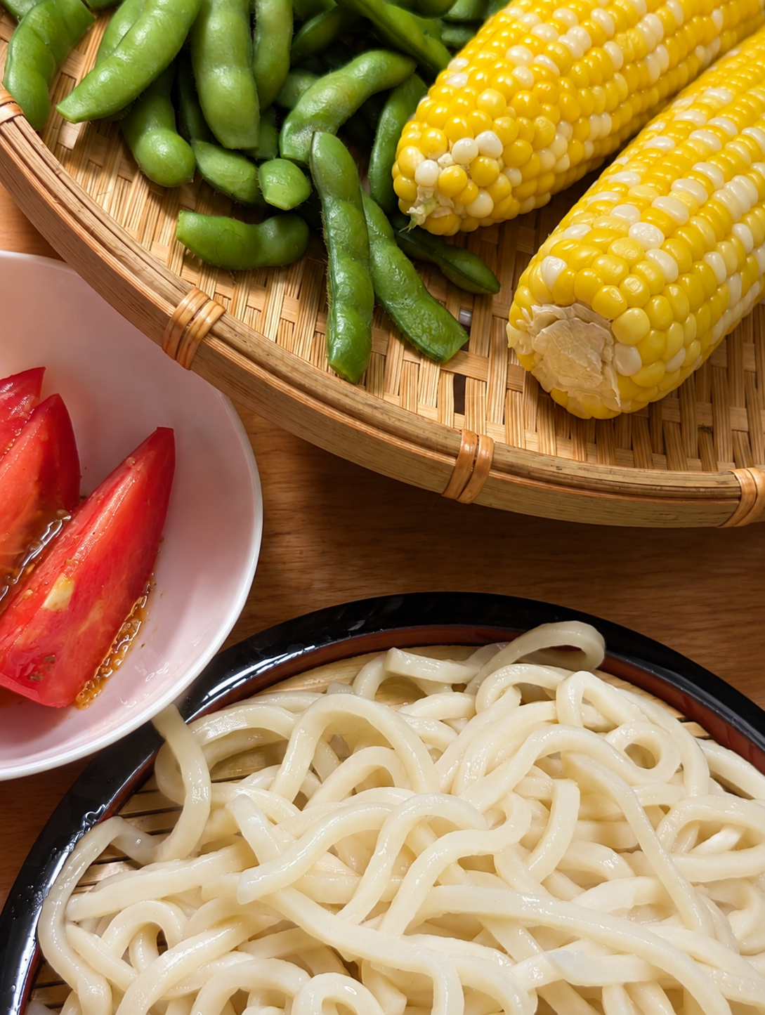 黒い器に盛られたざるうどんと、ざるに乗った枝豆とトウモロコシ、小鉢のトマト