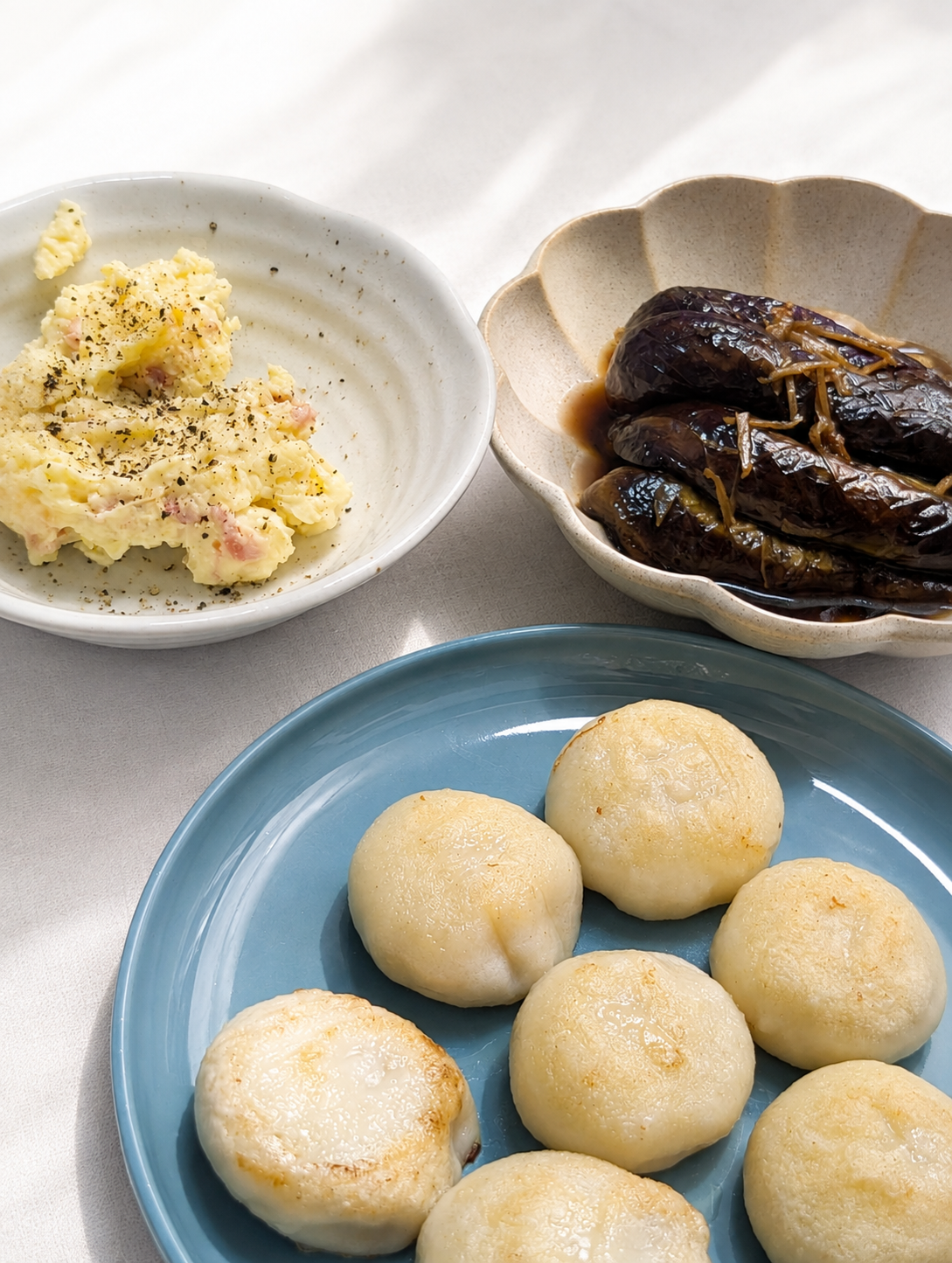 青い皿に並んだ丸い焼き餃子と、黒胡椒をかけたポテトサラダ、しょうがのせん切りをのせたなすの煮浸し