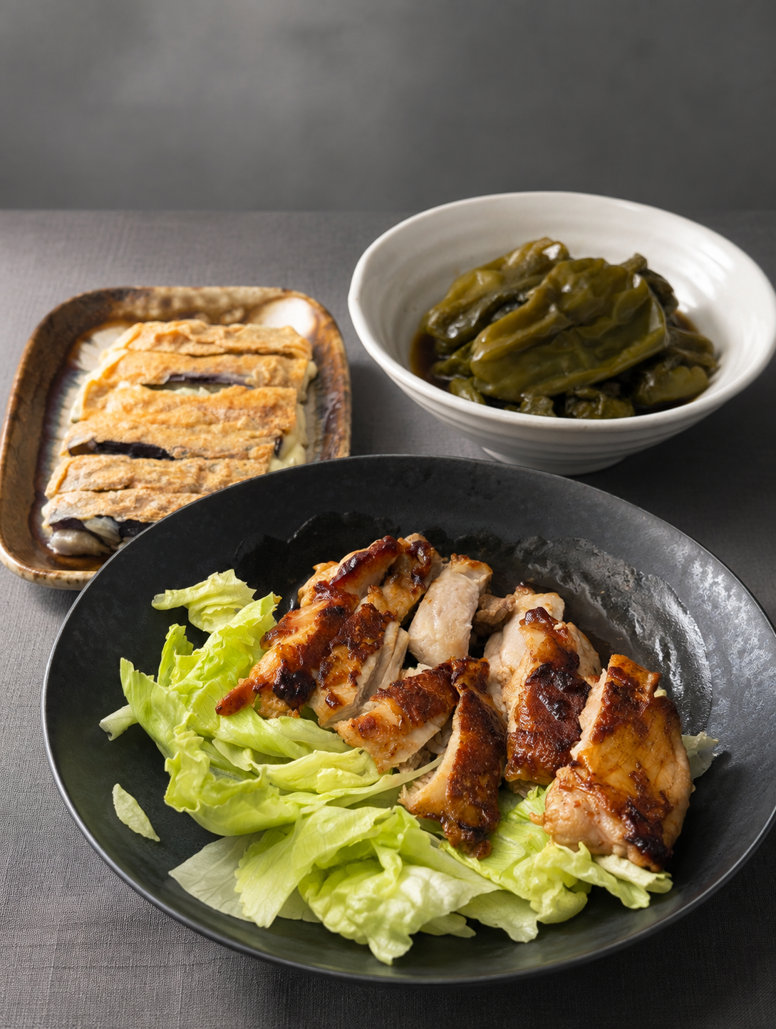 黒い皿にレタスを敷いて盛られた鶏の照り焼き。奥に白い小鉢のピーマンの煮浸しと、茄子のチーズはさみ