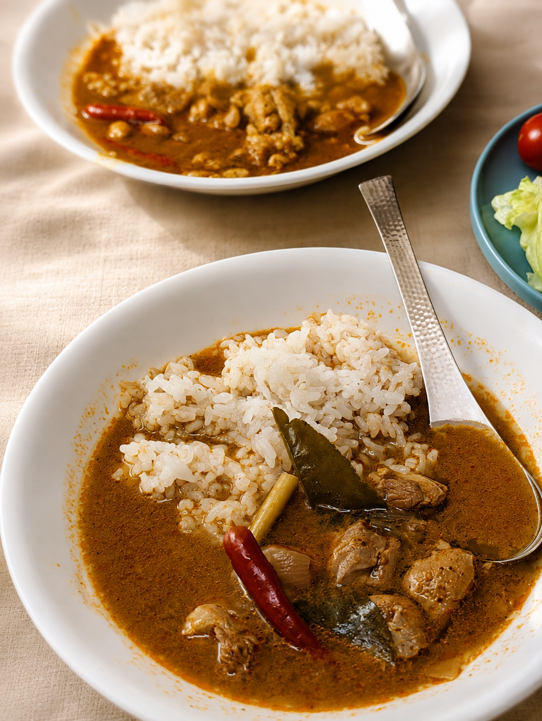 白い皿に盛られたタイカレー2皿。鶏肉と赤唐辛子とこぶみかんの葉が入り、副菜に白菜の漬物とミニトマト