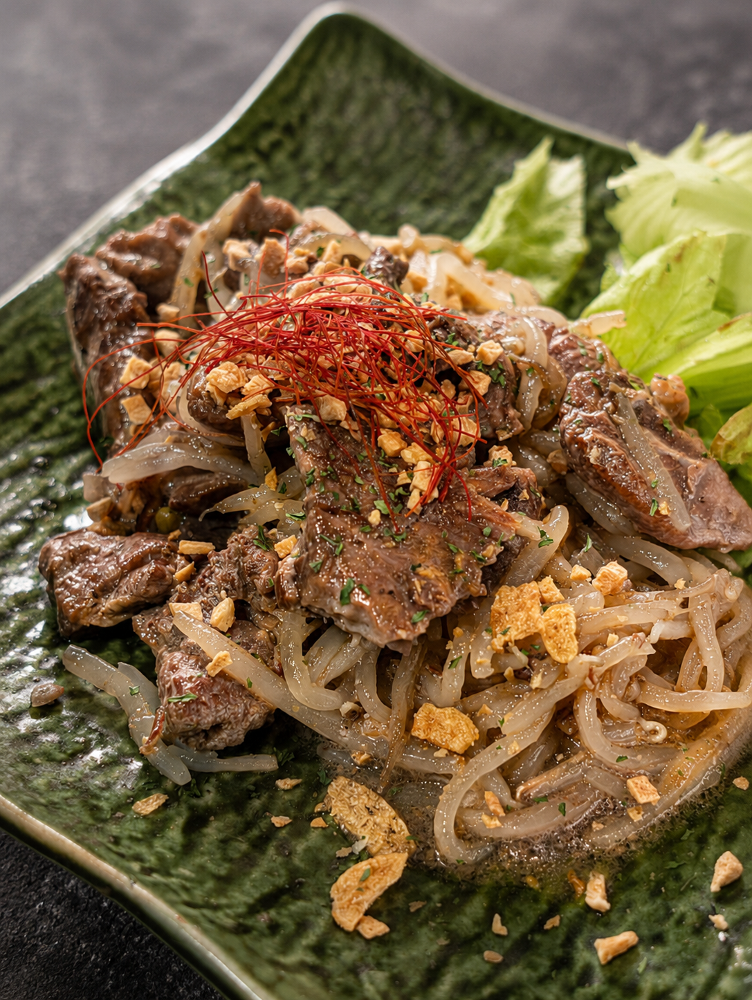 緑の皿に盛られた牛肉としらたきの炒め物。糸唐辛子とフライドガーリックがのっている