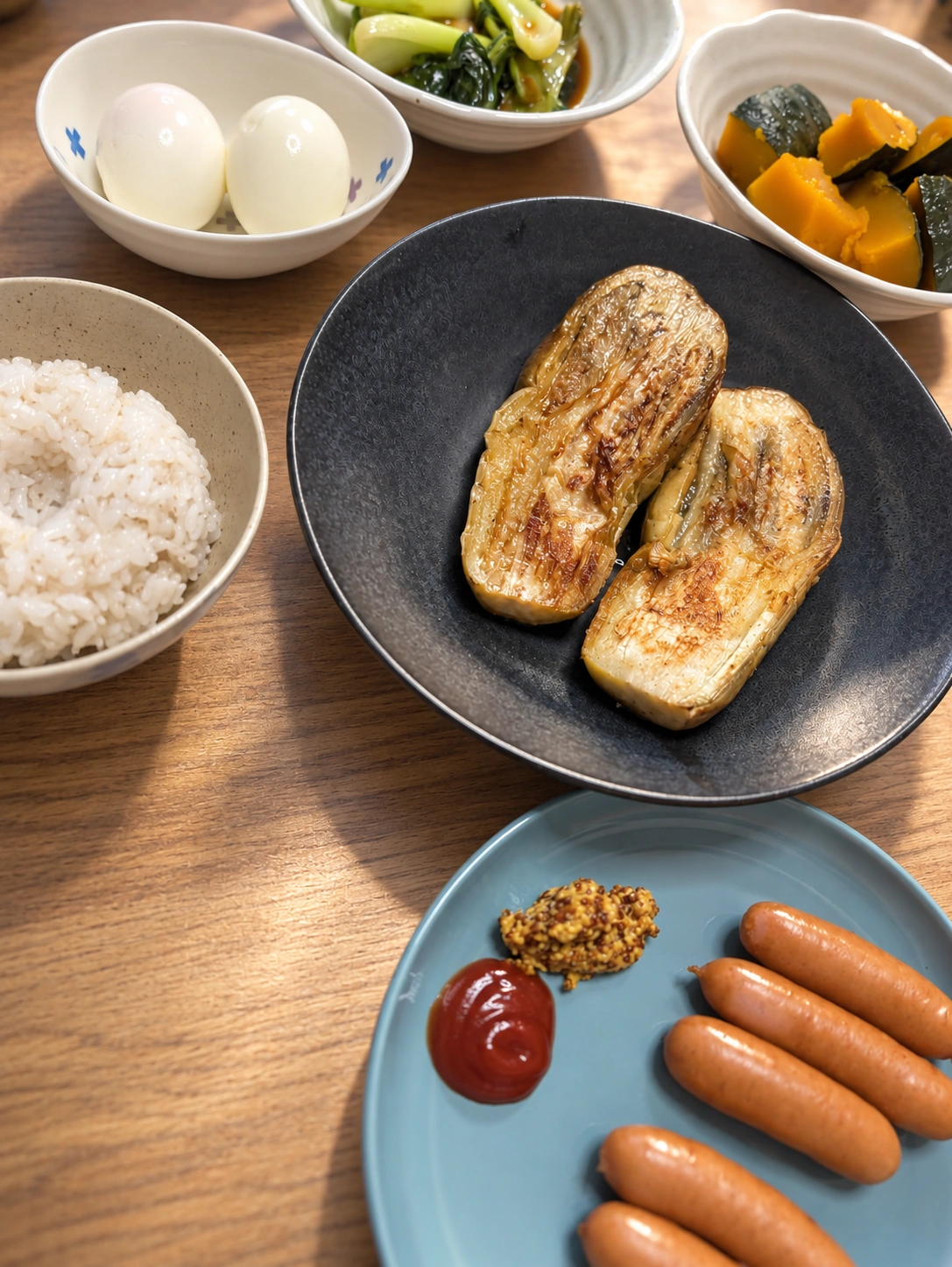 黒い皿の焼きナス2切れ、水色の皿のウインナーとケチャップと粒マスタード、ゆで卵、青梗菜の煮浸し、かぼちゃの煮物、ごはんを俯瞰で撮った食卓