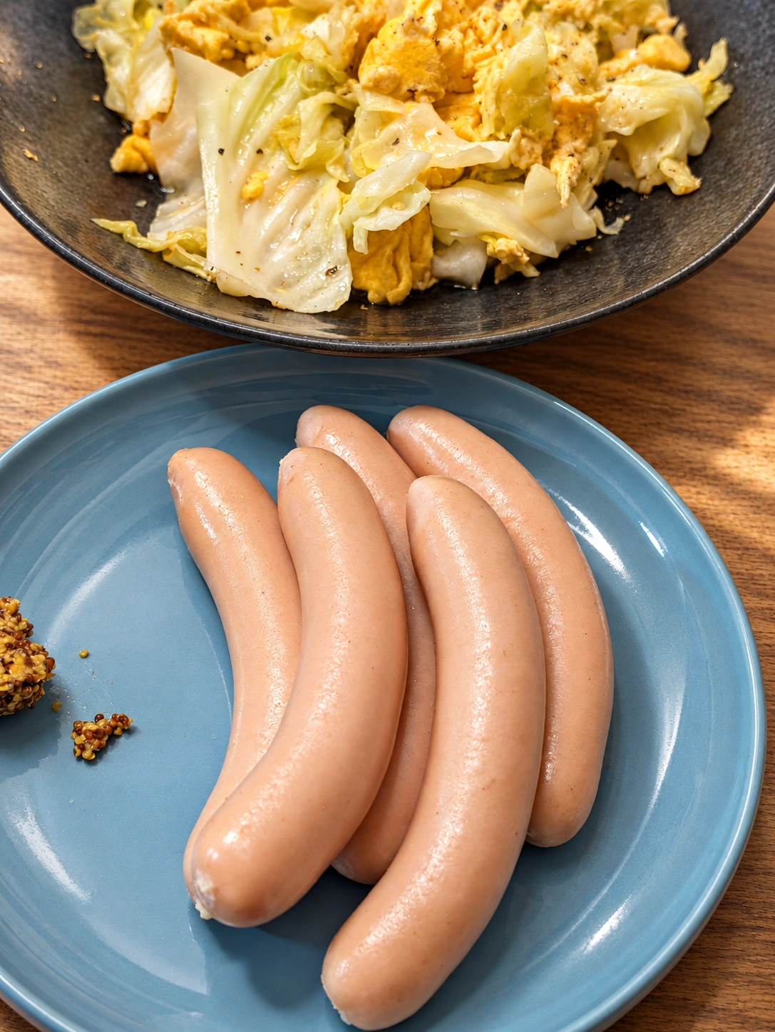 青い皿にスワンレイクビールのソーセージ4本と粒マスタード、奥にキャベツと卵の料理(卓上・光などをAIで加工)